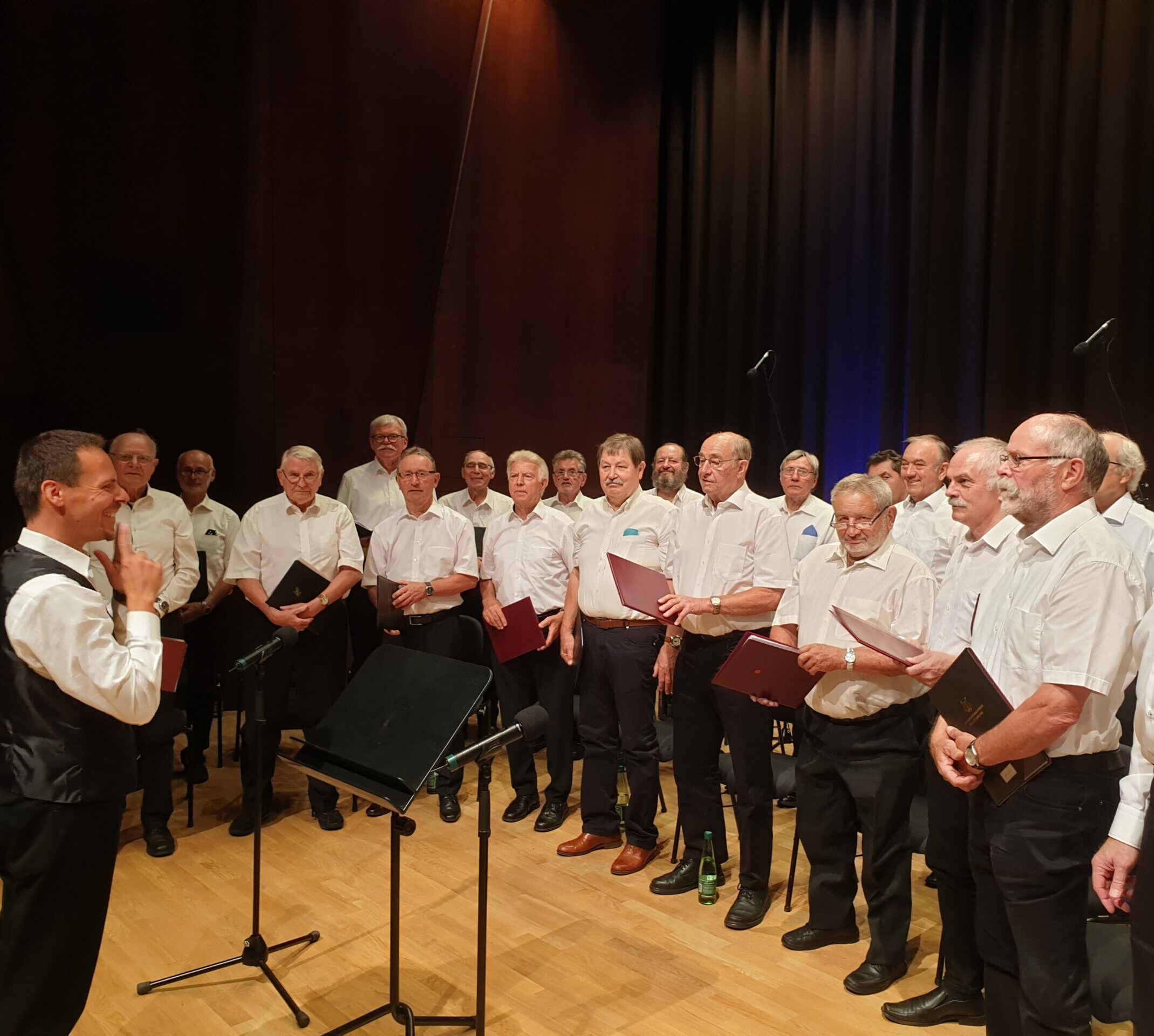 Der Männergesangsverein Kriegenbrunn-Falkendorf bereitet sich auf einen Auftritt vor. Viele Männer im weißen Hemd und schwarzer Hose stehen gemeinsam mit ihrem Chorleiter auf der Bühne, Chormappen in den Händen.